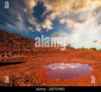 Riflesso delle nuvole in un piccolo puddle su secco suolo rosso al tramonto Foto Stock