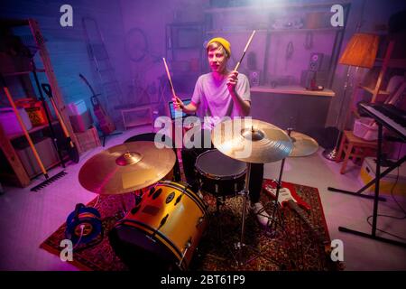 Giovane uomo in casualwear alzando le mani con i drumsticks mentre andando colpire i piatti durante le prove e la registrazione della musica in garage o in studio Foto Stock