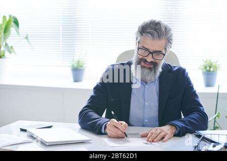 Professionista esperto in occhiali e formalwear che fa note su carta mentre si siede dalla scrivania contro la finestra e in attesa di clienti Foto Stock