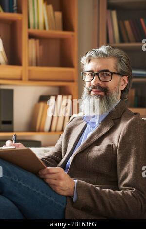 Psicologo esperto di occhiali e casualwear intelligente che ti guarda sullo sfondo di scaffali con letteratura professionale Foto Stock