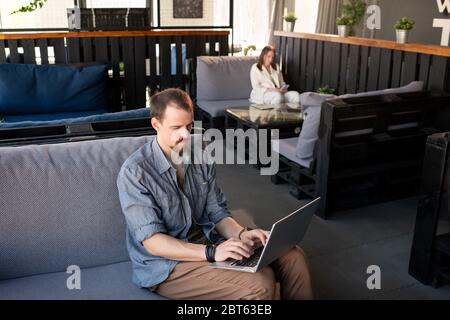 Giovane uomo occupato in casualwear seduto sul divano con il computer portatile in ginocchio e surf per negozi online che vendono attrezzature per ristoranti Foto Stock