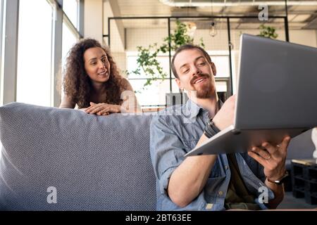 Giovane uomo sorridente portatore che mostra alla moglie nuove attrezzature per il loro ristorante mentre punta al display del laptop durante il lavoro Foto Stock