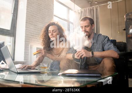 Giovane uomo contemporaneo che fuma la sigaretta elettronica mentre si siede accanto alla moglie che acquista online di fronte al computer portatile e usina carta di plastica Foto Stock