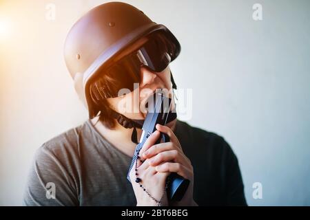 Un uomo vestito con equipaggiamento militare, un casco e occhiali protettivi, tiene una pistola aerosoft in mani e pranks, fingendo di mangiarla. Pericolo. Foto Stock