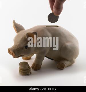 Mano che mette una moneta da 50 pence in una fessura di una banca di porcellini con una piccola pila di monete in primo piano Foto Stock