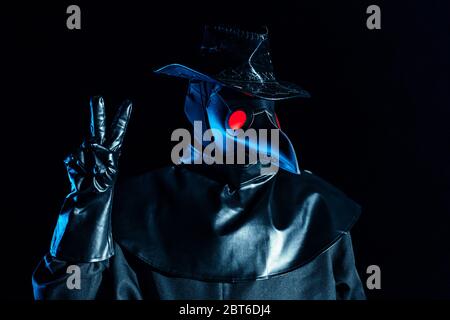 Uomo in costume medico peste con maschera corvo-come che mostra gesto di pace isolato su sfondo nero. Maschera creepy, concetto storico di costume. Epidemia Foto Stock