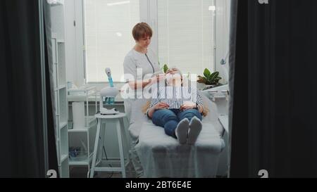 Cosmetologo applicazione maschera crema di fronte a bella donna presso salone di bellezza spa. Lei fa trattamenti anti-invecchiamento. Foto Stock