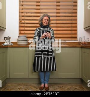 Donna anziana in piedi in cucina con grembiule a righe utilizzando il telefono cellulare Foto Stock