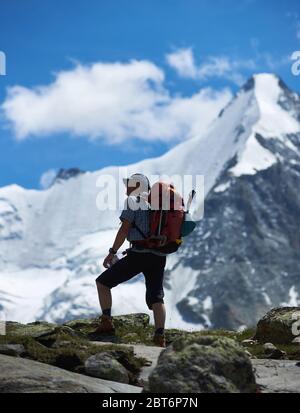 Zinal, Svizzera - 19 luglio 2019: Viaggiatore maschile con zaino che tiene una bottiglia d'acqua e guardando la vista delle colline innevate sotto il cielo blu. Turista in pantaloncini che si pausa durante l'escursione in montagna Foto Stock