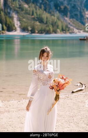 Bella sposa in abito bianco con maniche e pizzo, con bouquet giallo autunnale di fiori secchi e rose di pony, sul Lago di Braies in Italia Foto Stock