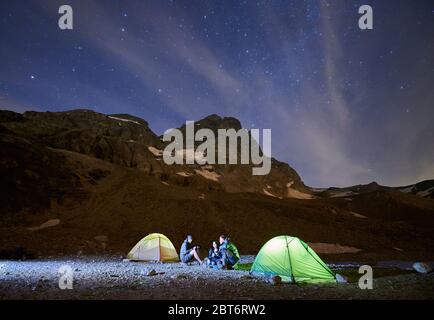 Magico scenario di cielo blu nuvoloso e stelle splendenti, bella zona di montagne. Tre turisti che chiacchierano, seduti vicino a tende illuminate. Splendida cresta di montagna con alte vette rocciose, paese delle meraviglie nelle Alpi Foto Stock