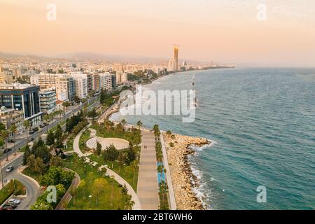 Vista aerea della costa cittadina di Limassol a Cipro. Passeggiata percorso Molos Park con palme, mare Mediterraneo e skyline urbano al tramonto, foto drone. Foto Stock