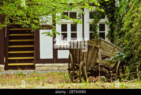 Una vecchia casa a telaio bianco, con scale sul lato sinistro e un vecchio carro di fieno in legno rotto sul lato destro Foto Stock