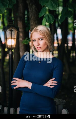 Ritratto di bella donna bionda in piedi davanti a piante verdi; lei indossa un elegante abito blu stretto. Ci sta guardando con le braccia incrociate Foto Stock