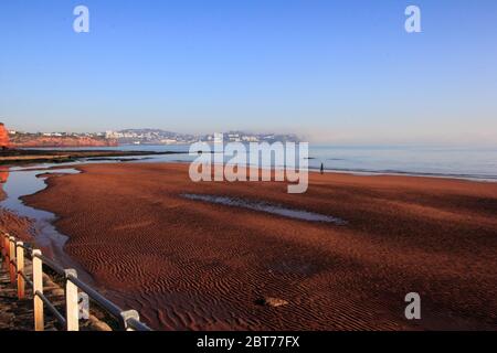 I Dog Walker sulla spiaggia di sabbia rossa increspata, Preston Sands, Paignton, Torbay con le scogliere di arenaria rossa che lavorano intorno a Torquay parzialmente avvolte in un mare Foto Stock