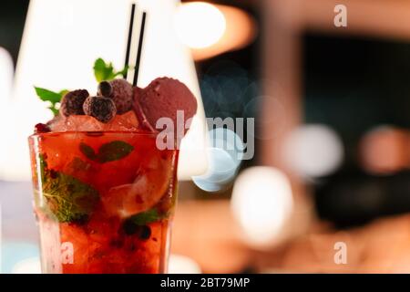 Mettere a fuoco in un bicchiere con un cocktail rosso decorato con frutti di bosco e gelato su un tavolo di notte Foto Stock