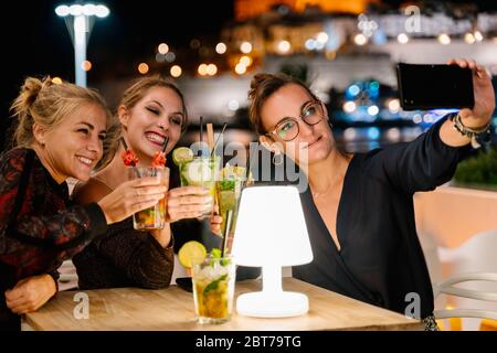 Tre ragazze che fanno un selfie mentre si siedono su una terrazza sorseggiando cocktail di notte con le luci della città sullo sfondo Foto Stock