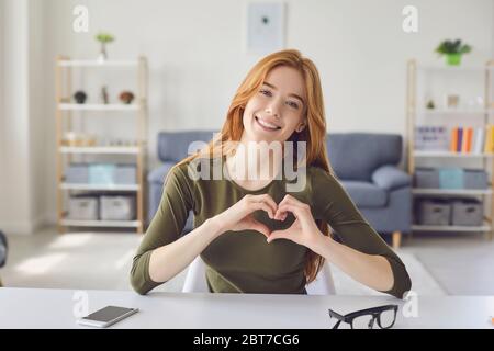Ottimista signora rossa sorridente per la macchina fotografica e gesturing cuore mentre si siede a tavolo in una stanza accogliente a casa Foto Stock