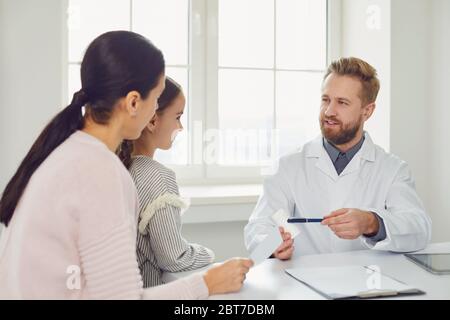 Medico maschile pediatrican dà a mamma e figlia una prescrizione, mentre seduto a un tavolo in un ufficio clinica. Foto Stock