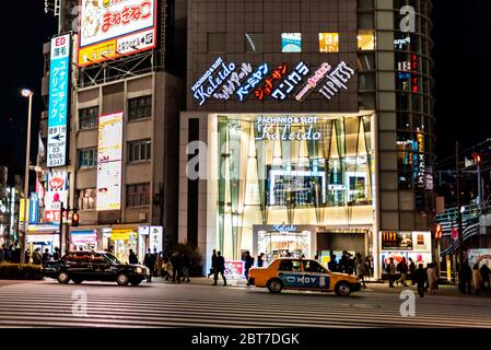 Shinjuku, Giappone - 3 aprile 2019: Via di Tokyo con traffico e persone che attraversano la strada in serata e ingresso pachinko slot machine casinò buildi Foto Stock