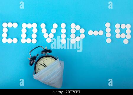 Iscrizione COVID-19 un da pillole mediche bianche e orologio nero di allarme con maschera medica protettiva si trova su uno sfondo di carta blu. Idea di attesa f Foto Stock