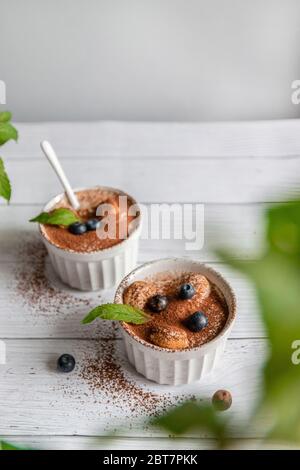 Classico dessert tiramisù con mirtilli, menta in vaso bianco su sfondo grigio. Concetto di posa piatta Foto Stock