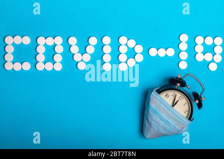 Iscrizione COVID-19 un da pillole mediche bianche e orologio nero di allarme con maschera medica protettiva si trova su uno sfondo di carta ciano. Idea in attesa Foto Stock