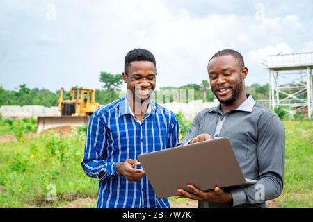 due imprenditori agricoli africani, che discutono di usare un computer portatile su un pezzo di terra Foto Stock
