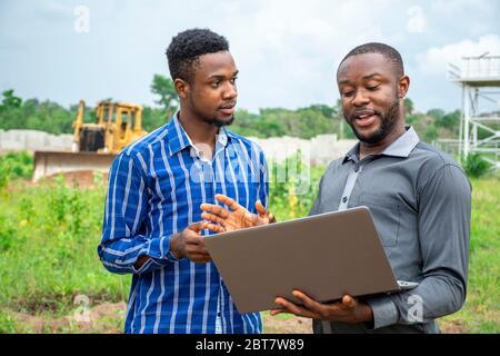 due imprenditori agricoli africani, che discutono di usare un computer portatile su un terreno Foto Stock