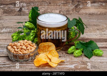 Un bicchiere di birra con arachidi e patatine. Giro verde con coni. Bevanda schiumosa. Ancora vita su sfondo di legno Foto Stock