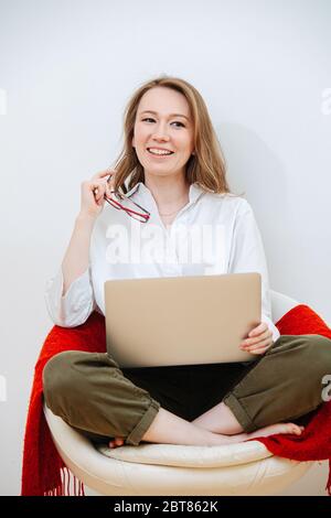 Sorridente ragazza lavora a casa con un computer portatile seduto in lotus posa su una sedia Foto Stock