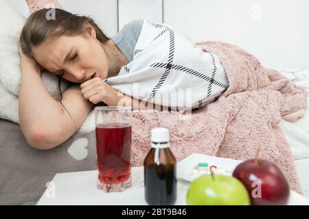 L'adolescente tosse. Gli intasa la bocca con la mano. Si trova sul letto. Frutta e medicina sul tavolo. Foto Stock