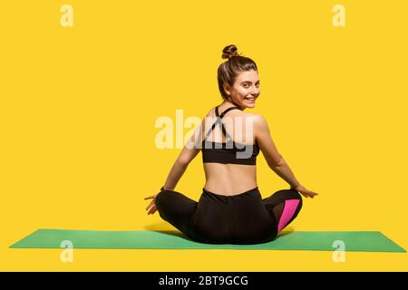 Indietro vista allegra donna con capelli bun in stretto abbigliamento sportivo seduta in posizione lotus sulla palestra mat praticare yoga, girando testa a macchina fotografica e sorridente jo Foto Stock