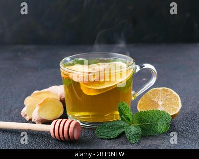 Tè alle erbe con zenzero, menta, limone e miele. Tazza di tè caldo con vapore e ingredienti su fondo nero di cemento. Spazio di copia Foto Stock