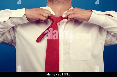 l'uomo adulto in una camicia bianca dell'ufficio strappa un filo rosso satinato dal collo su uno sfondo blu, concetto di depressione, disoccupazione e licenziamento Foto Stock