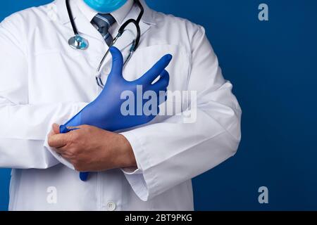 il medico maschile in uniforme bianca mette sulle sue mani guanti di lattice sterili blu, sfondo blu, spazio per la copia Foto Stock