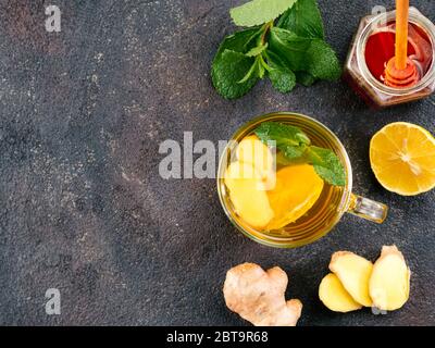 Tè alle erbe con zenzero, menta, limone e miele. Tazza di tè caldo con vapore e ingredienti su fondo nero di cemento. Spazio di copia. Vista dall'alto o piatta. Foto Stock