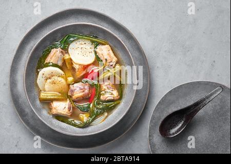 Zuppa di carne di maiale Sinigang na Baboy o Filippino in ciotola grigia su fondo di cemento. Sinigang è un piatto di cucina filippina con carne, bamia, daikon, spinaci, Foto Stock