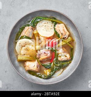 Zuppa di carne di maiale Sinigang na Baboy o Filippino in ciotola grigia su fondo di cemento. Sinigang è un piatto di cucina filippina con carne, bamia, daikon, spinaci, Foto Stock