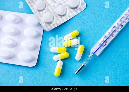 termometro analogico su fondo blu con gruppo di medicamenti in capsule bianche/gialle, covid19, virus della corona, Foto Stock