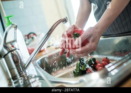 Le mani femminili lavano il pepe rosso sotto un flusso d'acqua, sullo sfondo delle verdure nel lavello. Cucina cibo. Foto Stock