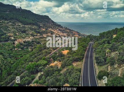 Fotografia aerea drone punto di vista vuoto ondeggiante strada asfaltata attraverso alberi lussureggianti piccolo villaggio di Banyalbufar, cielo nuvoloso moody sotto Mediterraneo Foto Stock