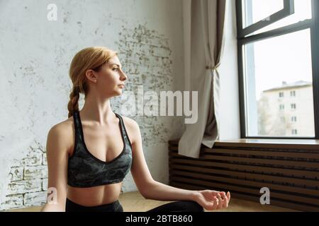 Giovane donna sana e bella seduta a casa in posizione yoga. Una ragazza bella è impegnata nella meditazione a casa in un luminoso interno. Foto Stock