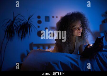 Ritratto di giovane donna riccia con smartphone e sorridente felice mentre si sdraia sul letto in camera oscura, copia spazio Foto Stock