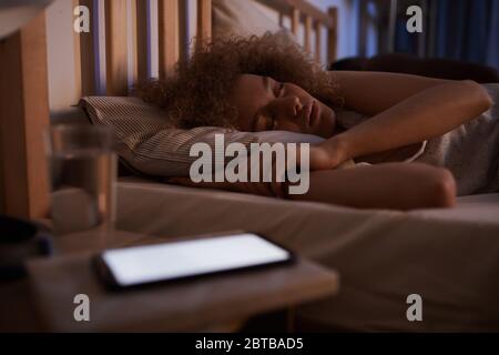 Ritratto di giovane donna dai capelli ricci che dorme tranquillamente a letto di notte con smartphone illuminato sul comodino, spazio per la copia