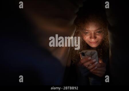 Ritratto di giovane donna con capelli ricci che guarda lo schermo dello smartphone e sorride mentre si sdraia a letto in camera oscura, copia spazio Foto Stock