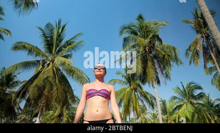 gioiosa bruna ragazza in bikini cammina lungo palmeti contro il cielo blu godendo di natura esotica sotto la luce del sole basso angolo shot Foto Stock