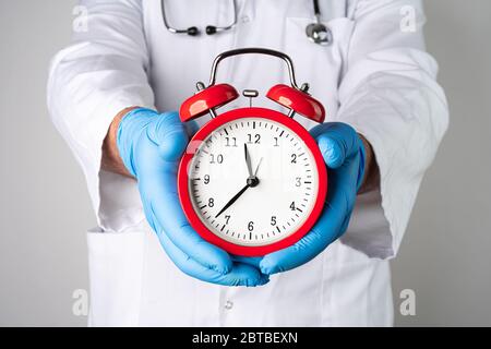 Uomo caucasico in posa come medico che tiene in mano un orologio rosso vintage allarme in guanti protettivi. Tempo di fine quarantena o concetto astratto pandemico. Foto Stock