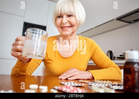 Pensione. Donna anziana seduta al tavolo in cucina tenendo una tazza d'acqua con il trattamento di influenza di dissolvere una compressa sorridente felice Foto Stock