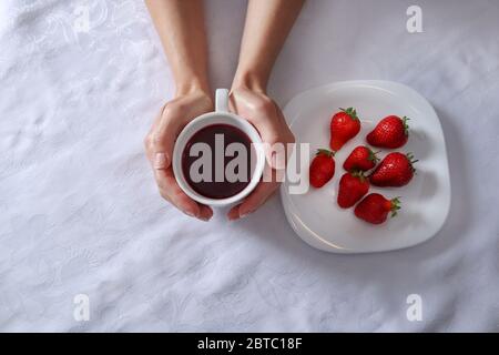 Donna mano che tiene una tazza di tè accanto ad un piatto pieno di fragole frutta. Un piatto e una ragazza mani su una tovaglia bianca Foto Stock